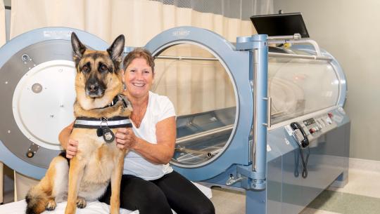 Patient sits in front of the hyperbaric oxygen chamber with her german shepherd