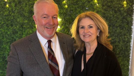 Bob and Cathy Creighton stand in front of an ivy wall with string lights