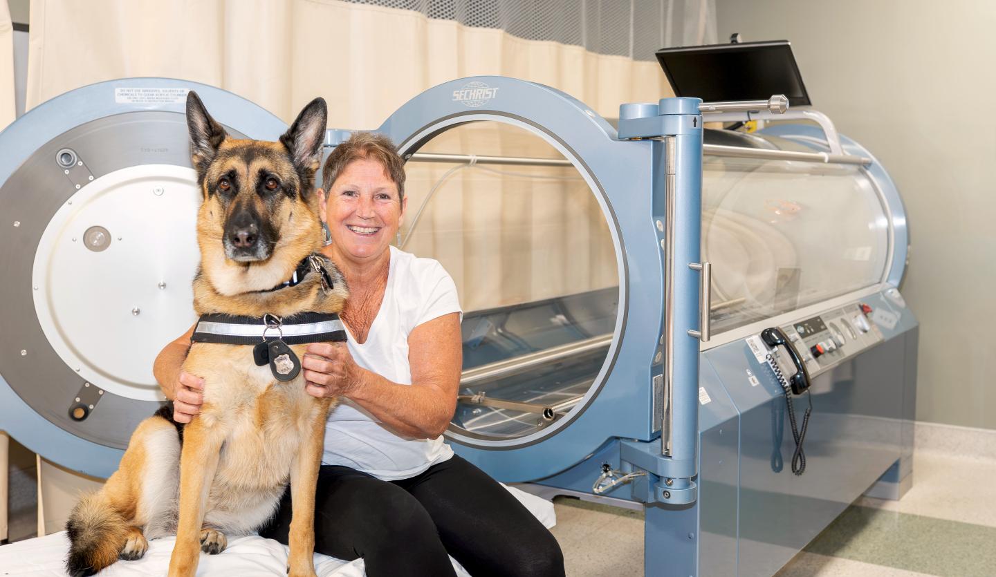 Patient sits in front of the hyperbaric oxygen chamber with her german shepherd